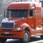 Freightliner Columbia semi truck in orange, hauling logs, showcases headlight design shared with Mercedes-Benz ML-Class.