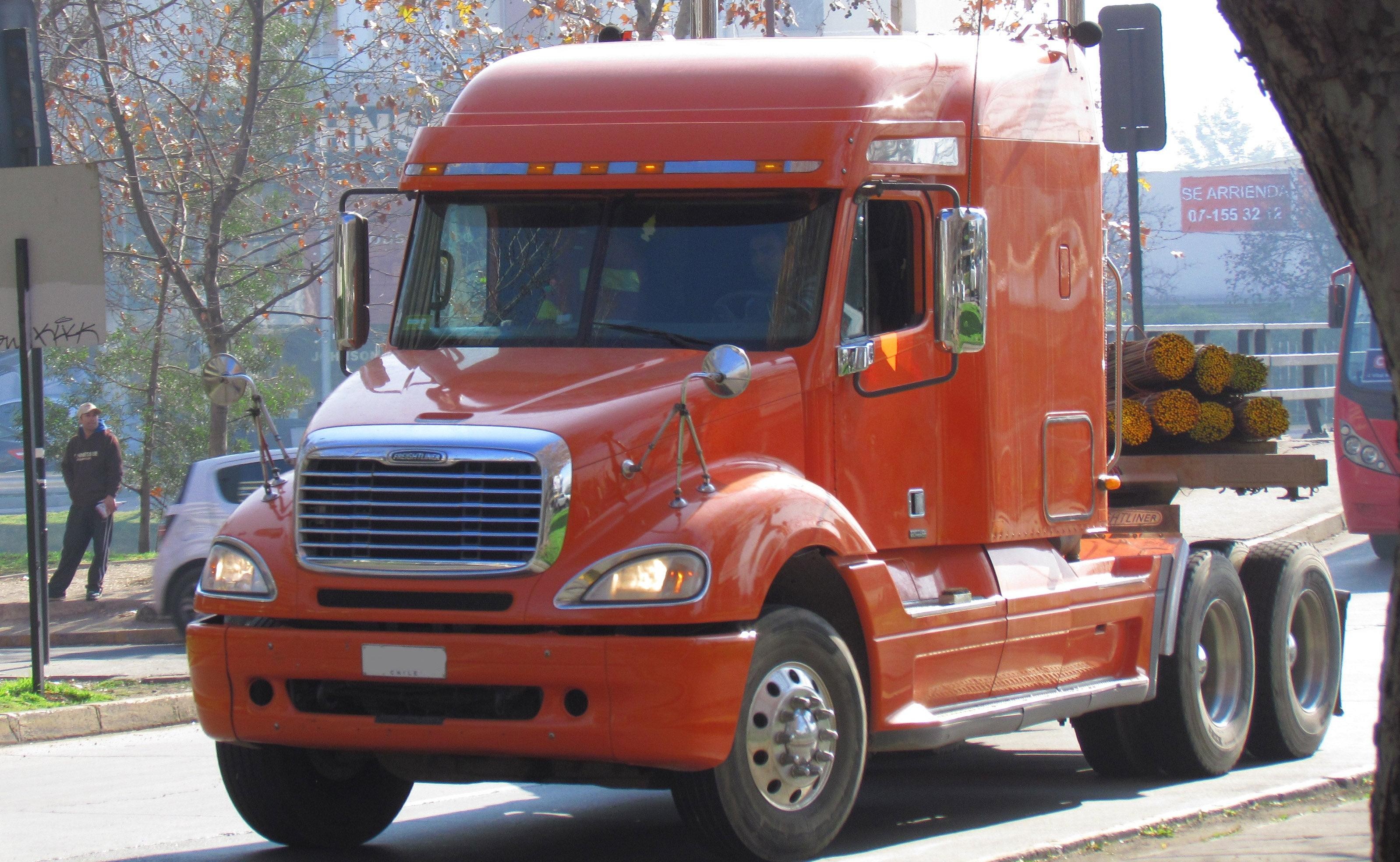 Freightliner Columbia semi truck in orange, hauling logs, showcases headlight design shared with Mercedes-Benz ML-Class.